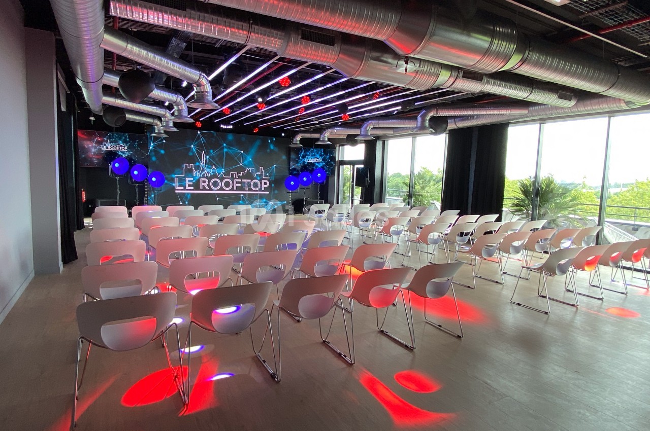 Salle de conférence moderne avec rangées de chaises blanches, éclairage coloré et grande baie vitrée donnant sur l'extérieur.