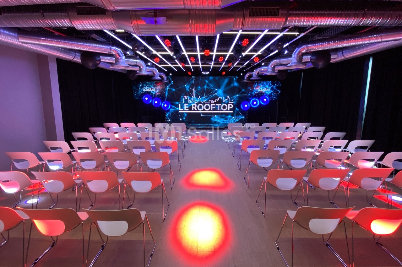 Salle de conférence moderne avec rangées de chaises blanches, éclairages colorés et scène équipée d'un écran LED.