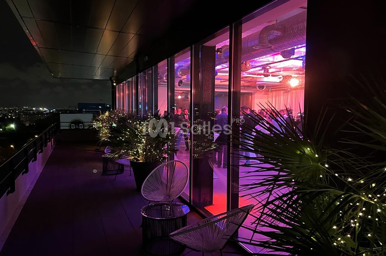 Vue nocturne d'une terrasse illuminée avec des plantes et des fauteuils, donnant sur une salle éclairée aux tons colorés.