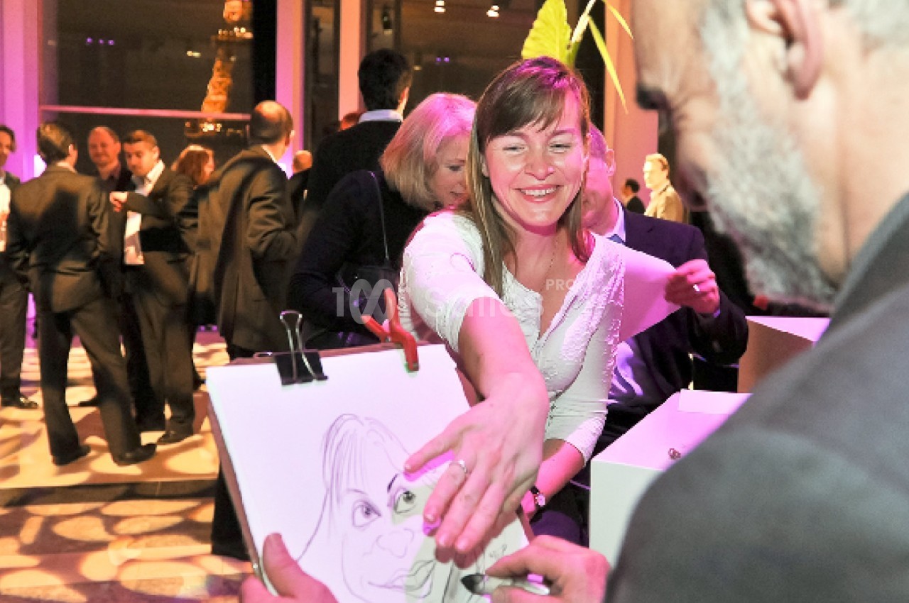Une femme souriante récupère un dessin caricatural lors d'un événement avec des invités en arrière-plan.