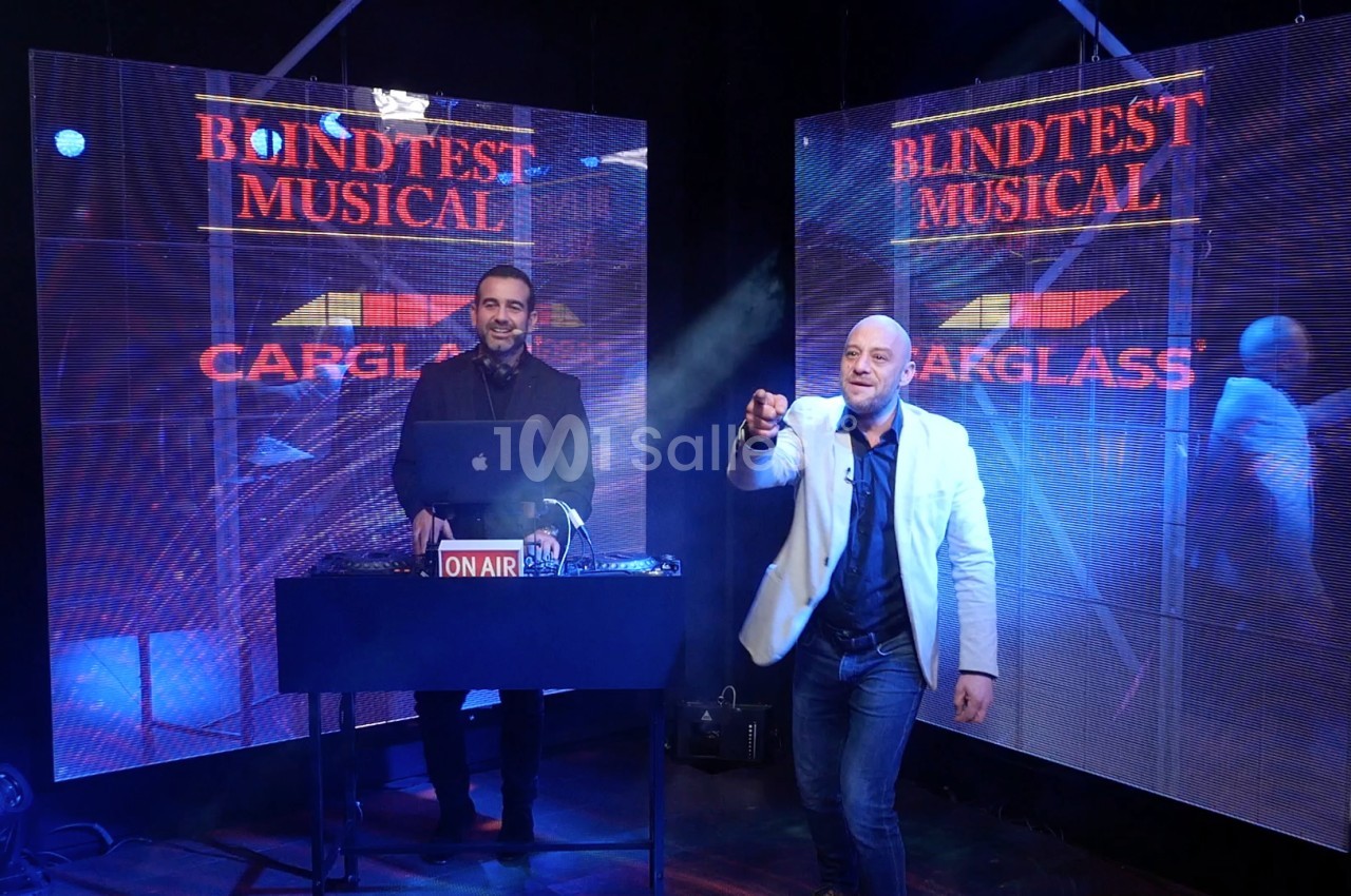 Deux hommes sur une scène avec un DJ, éclairages colorés et écrans affichant ’Blindtest Musical Carglass’.