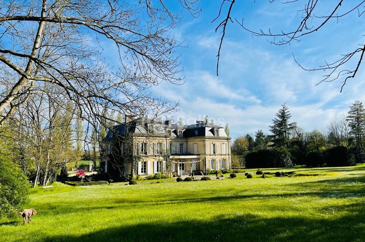 Manoir en pierre entouré d'un grand jardin verdoyant sous un ciel bleu clair.