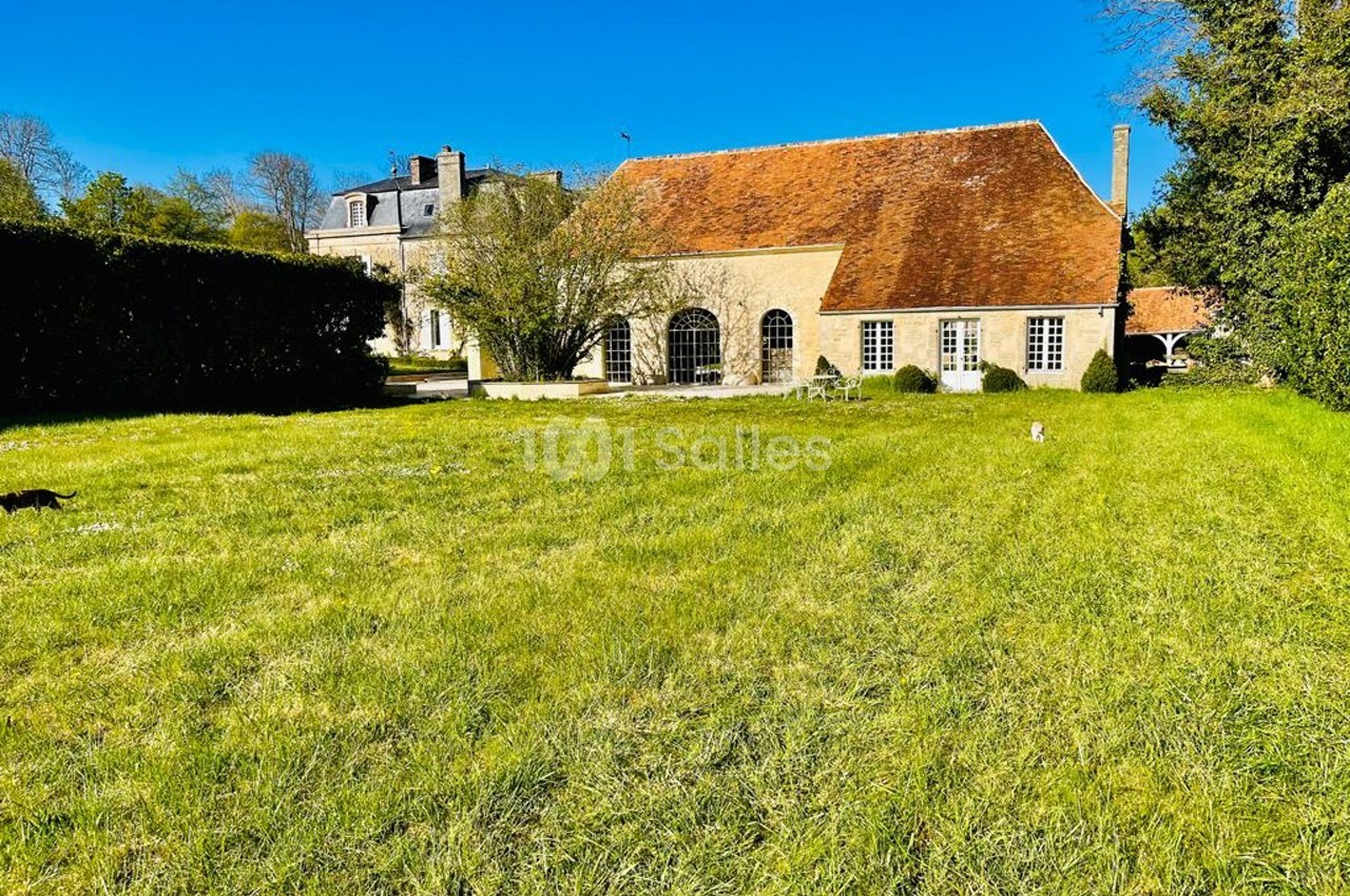 Grande maison en pierre avec toit en tuiles rouges, entourée d'un grand jardin verdoyant sous un ciel bleu.