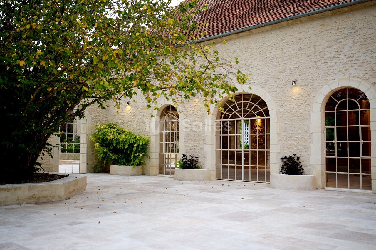Cour pavée avec un arbre, des plantes en pots et une façade en pierre ornée de grandes fenêtres cintrées.