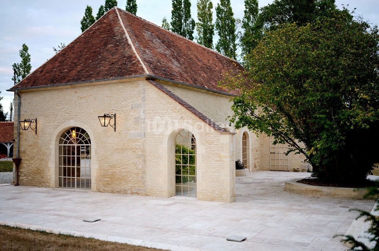 Bâtiment en pierre avec toit en tuiles rouges, grandes fenêtres en arc et terrasse pavée entourée de verdure.