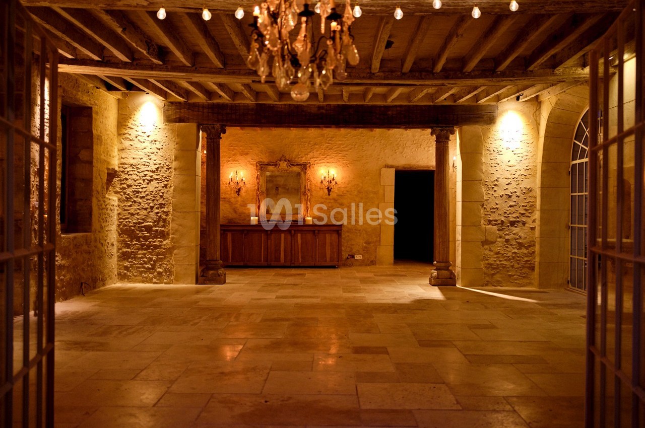 Salle en pierre avec un sol en dalles, un lustre suspendu, des murs éclairés et un meuble en bois au fond.