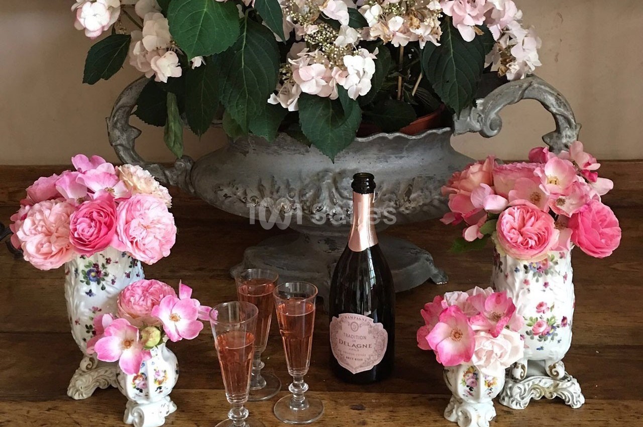 Vase ancien et flûtes de champagne rosé entourés de bouquets de fleurs roses sur une table en bois.