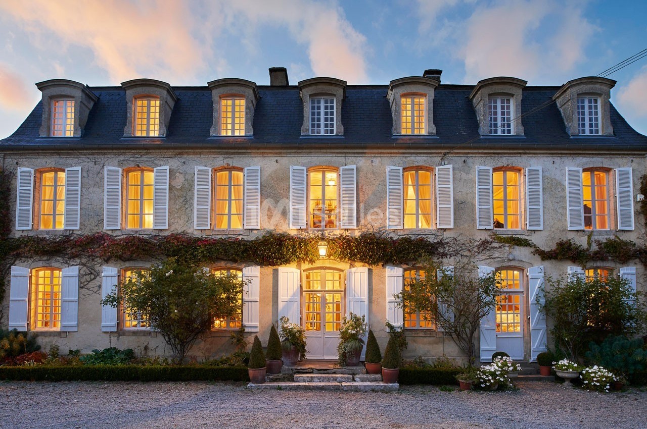 Façade d'une grande maison en pierre avec volets blancs, éclairée de l'intérieur au crépuscule.