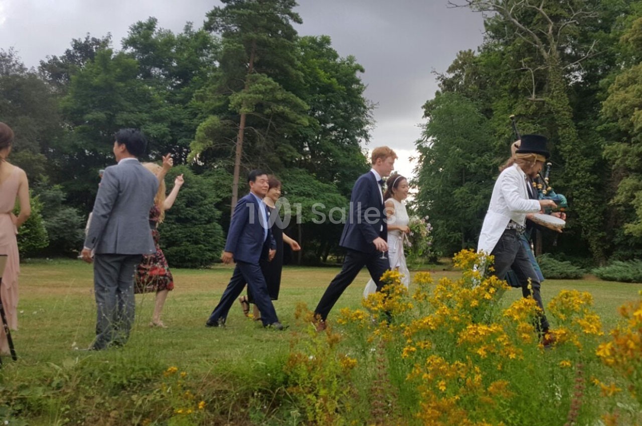 Un groupe de personnes marche dans un parc verdoyant, avec un homme en costume et chapeau haut-de-forme en tête.