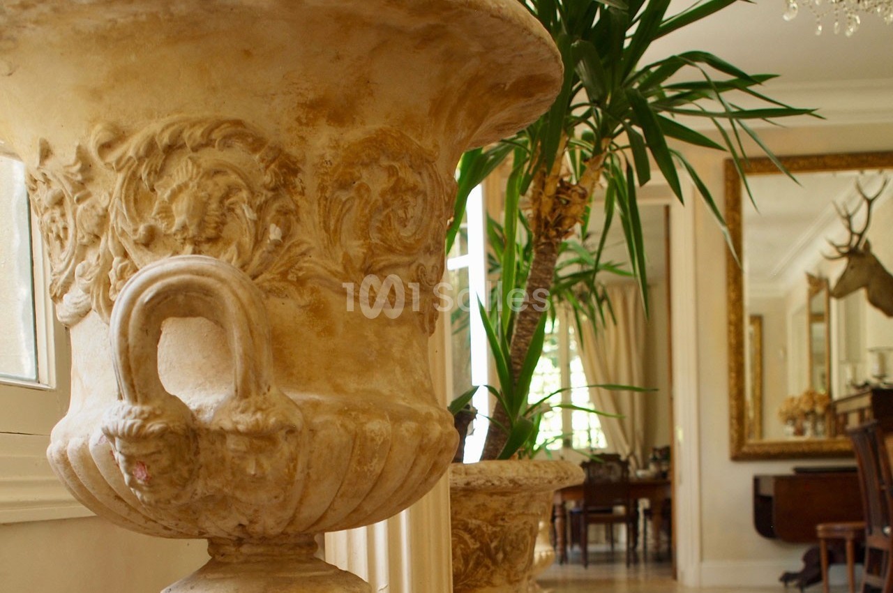 Grand vase décoratif en pierre sculptée devant un intérieur élégant avec miroir doré et plantes.