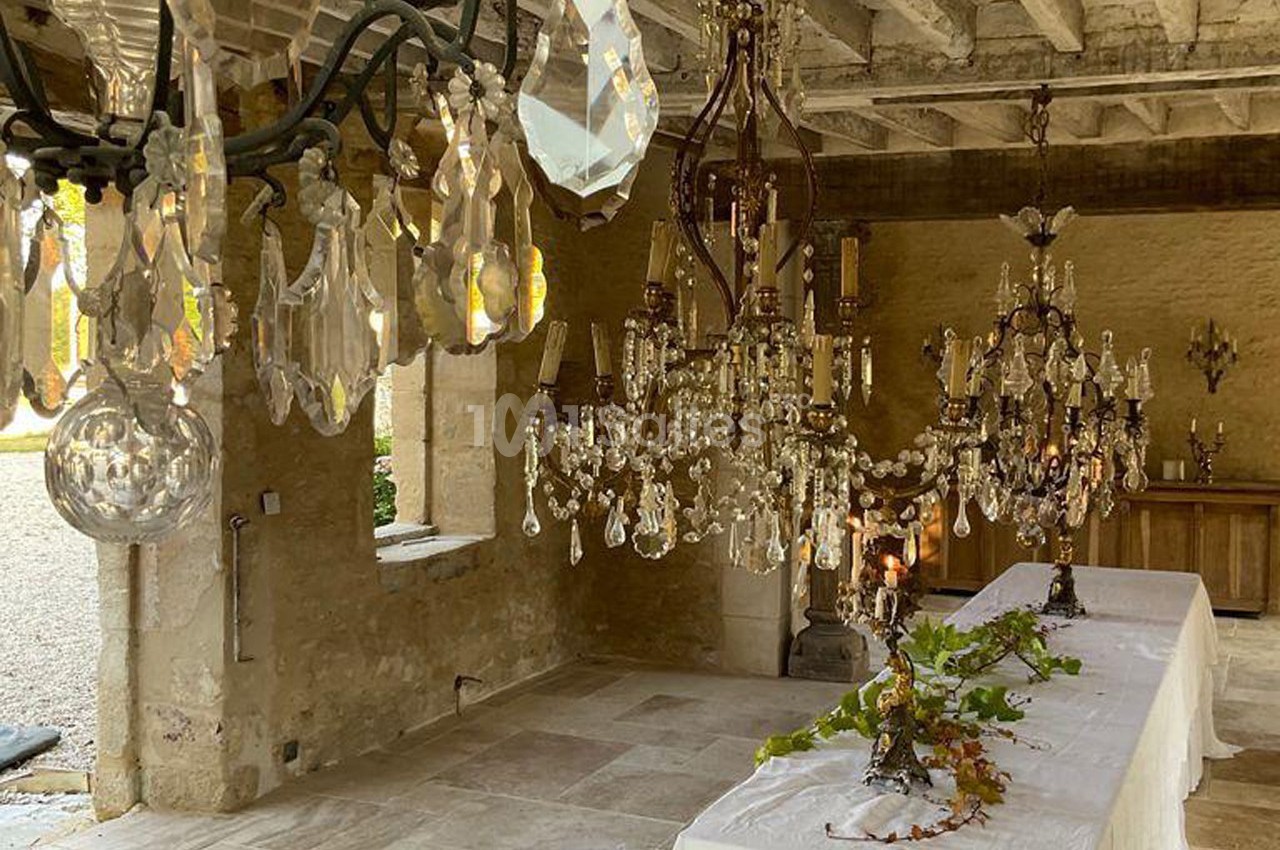 Des lustres en cristal suspendus éclairent une salle rustique avec des tables décorées de nappes blanches et de feuillage.