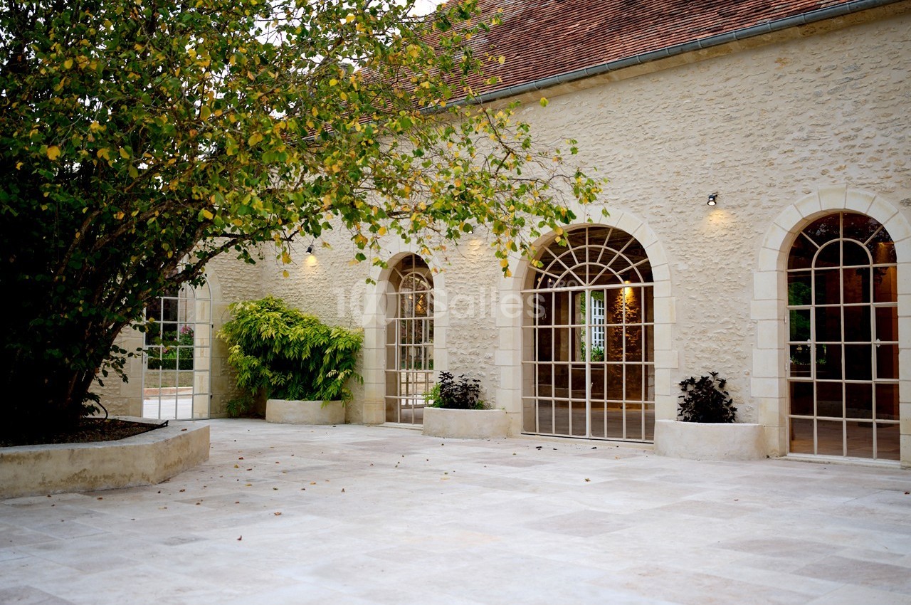 Cour pavée avec un arbre, des plantes en pots et une façade en pierre ornée de grandes fenêtres cintrées.