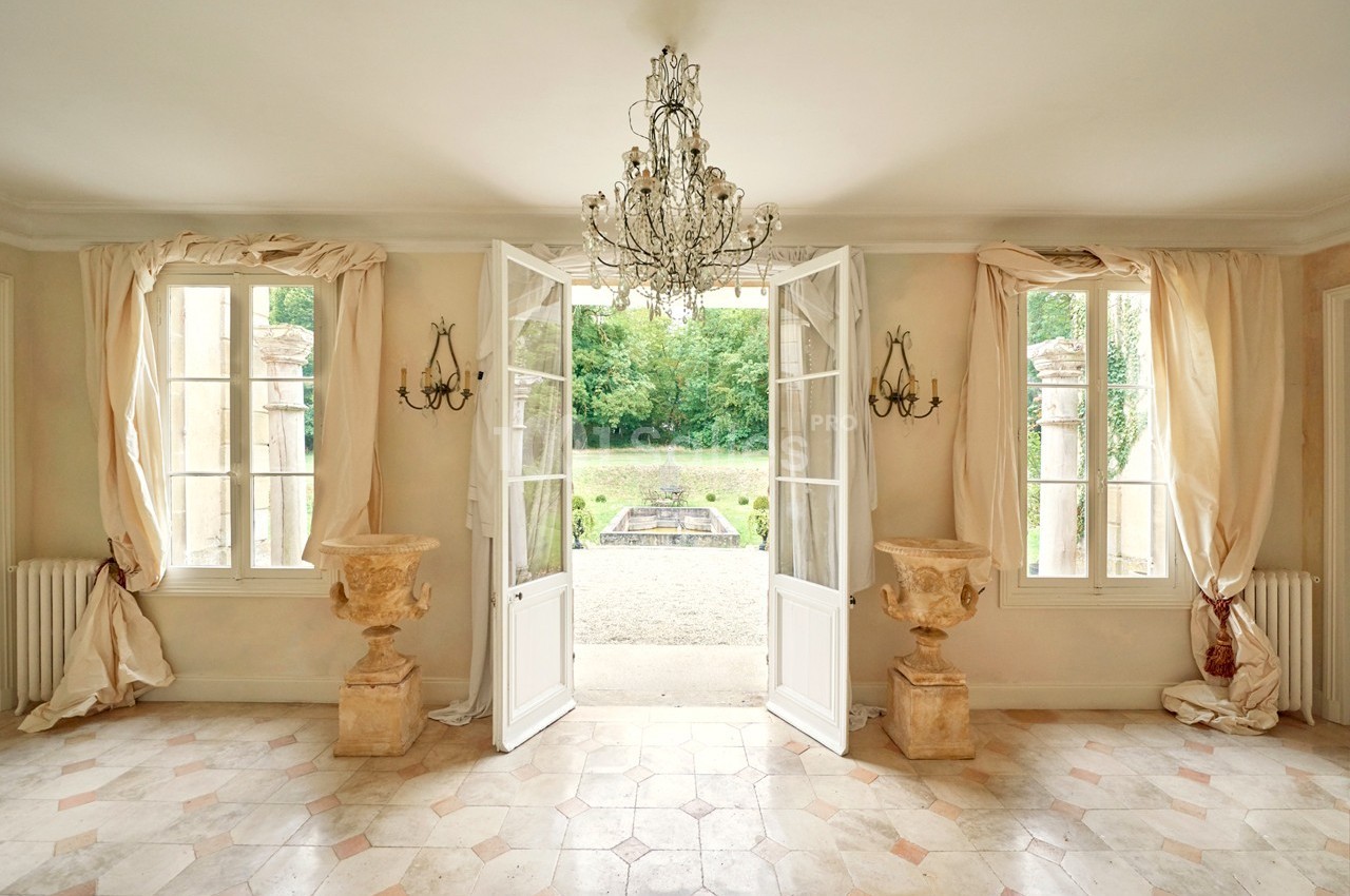 Salle lumineuse avec portes-fenêtres ouvertes sur un jardin, lustre central et décorations en pierre.