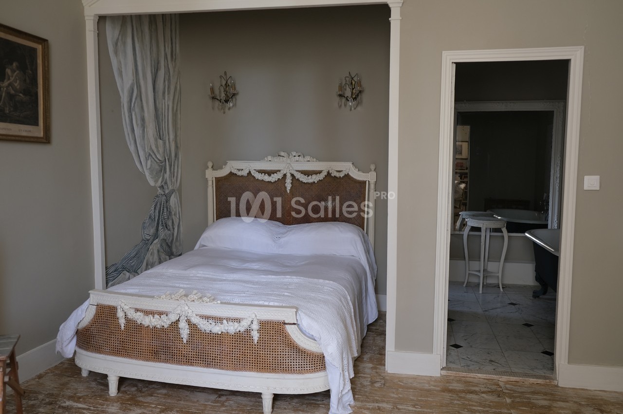 Lit double avec tête de lit en rotin et draperies, dans une chambre au sol en bois, avec une ouverture vers une autre pièce.