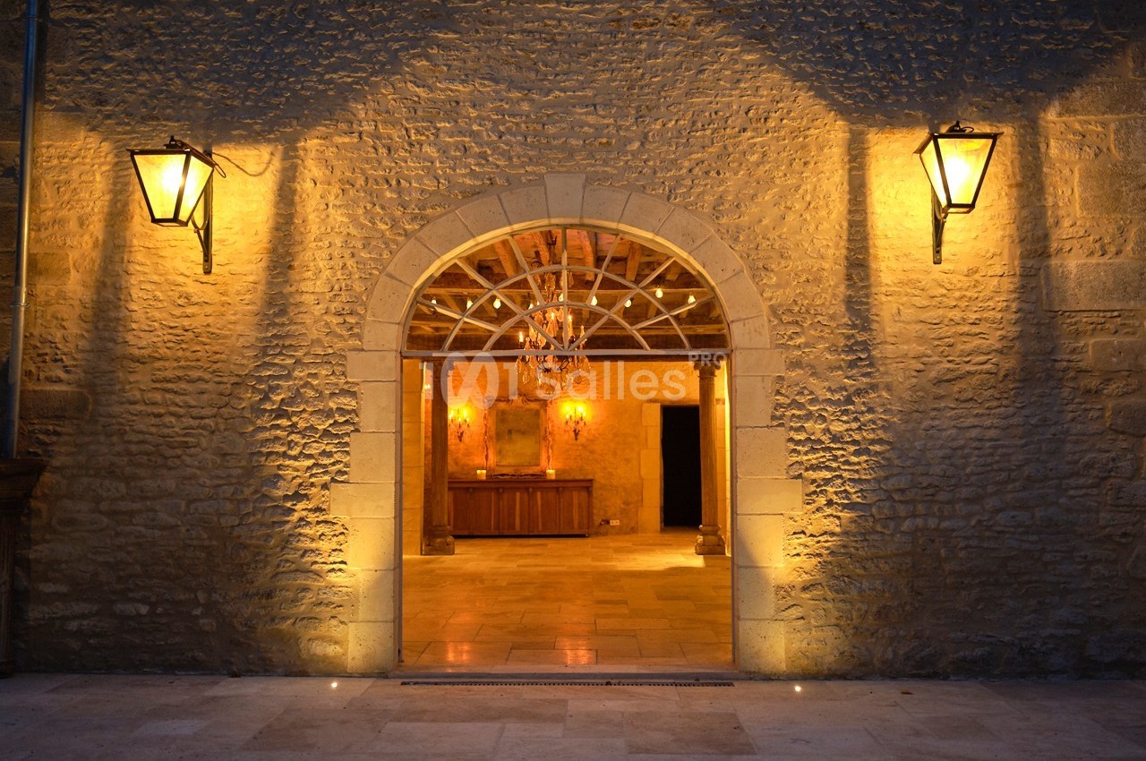 Entrée voûtée en pierre éclairée par des lanternes, donnant sur une salle avec un lustre et des murs en pierre.