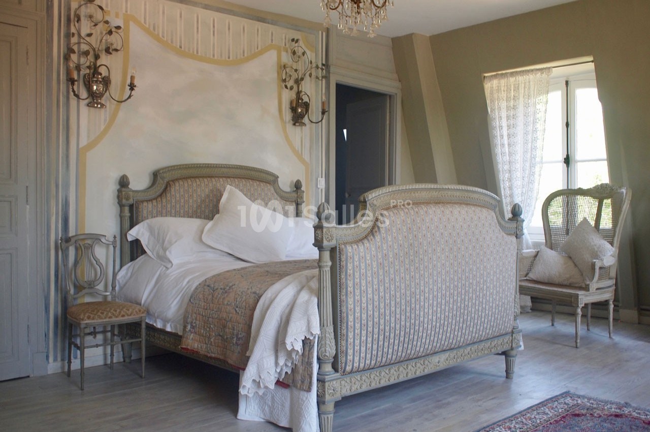 Chambre lumineuse avec un lit double ancien, des murs peints, un lustre et des fauteuils près d'une fenêtre.