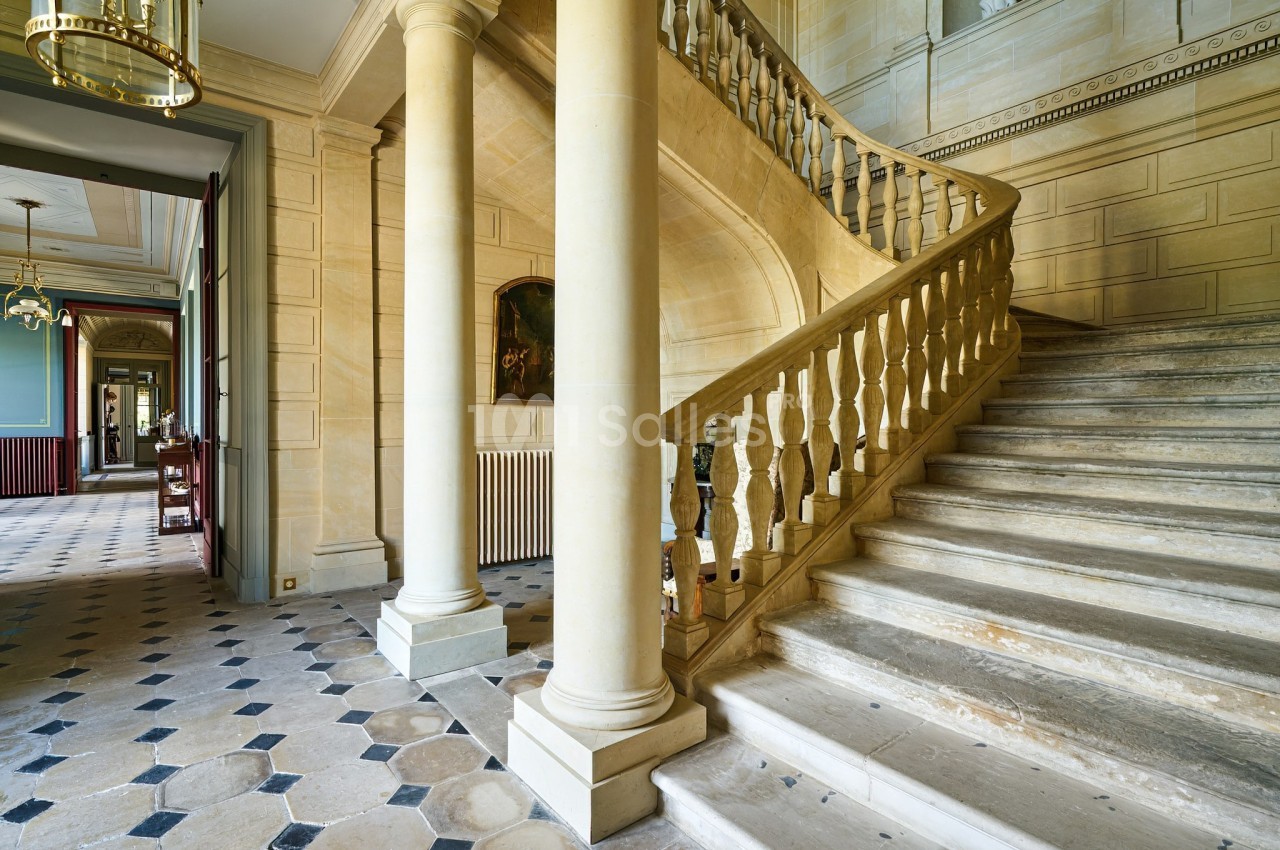 Escalier en pierre avec balustrade sculptée dans un intérieur lumineux aux murs clairs et sol en carreaux.