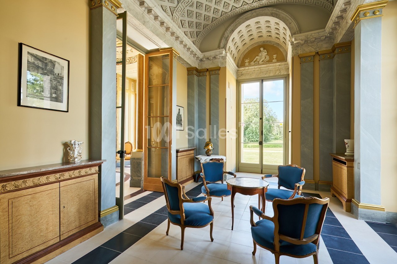 Salle élégante avec mobilier ancien, chaises bleues, table centrale, grandes fenêtres et décorations classiques.