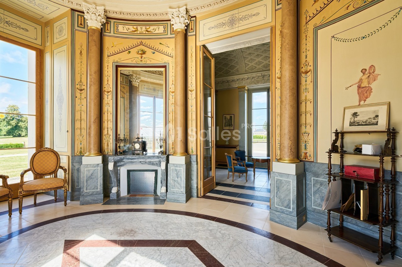 Intérieur d'une pièce élégante avec colonnes dorées, cheminée, miroirs et vue sur un jardin à travers de grandes fenêtres.