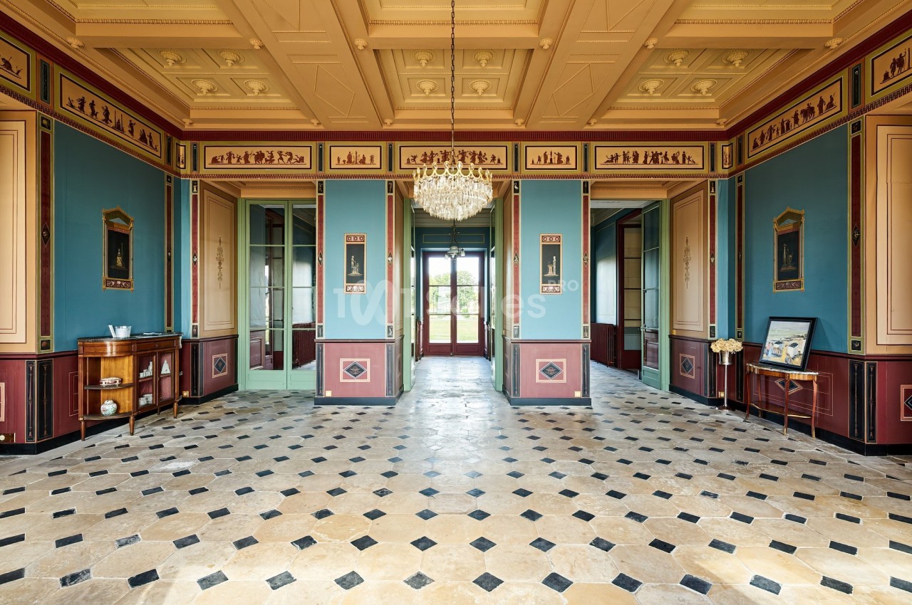 Salle élégante avec un sol en damier, murs ornés de moulures et peintures, et un lustre central suspendu.