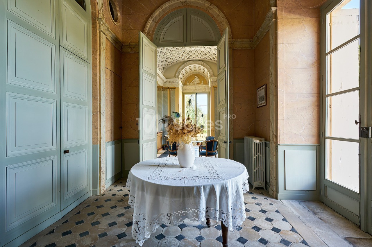Intérieur lumineux avec table ronde ornée d'une nappe blanche et vase de fleurs séchées, portes ouvertes sur un salon.