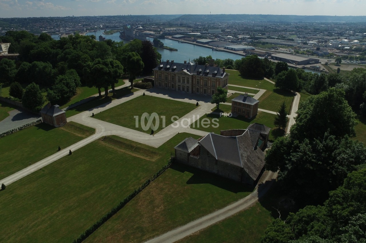 Vue aérienne d’un château entouré de jardins et de pelouses, avec une rivière et une ville en arrière-plan.