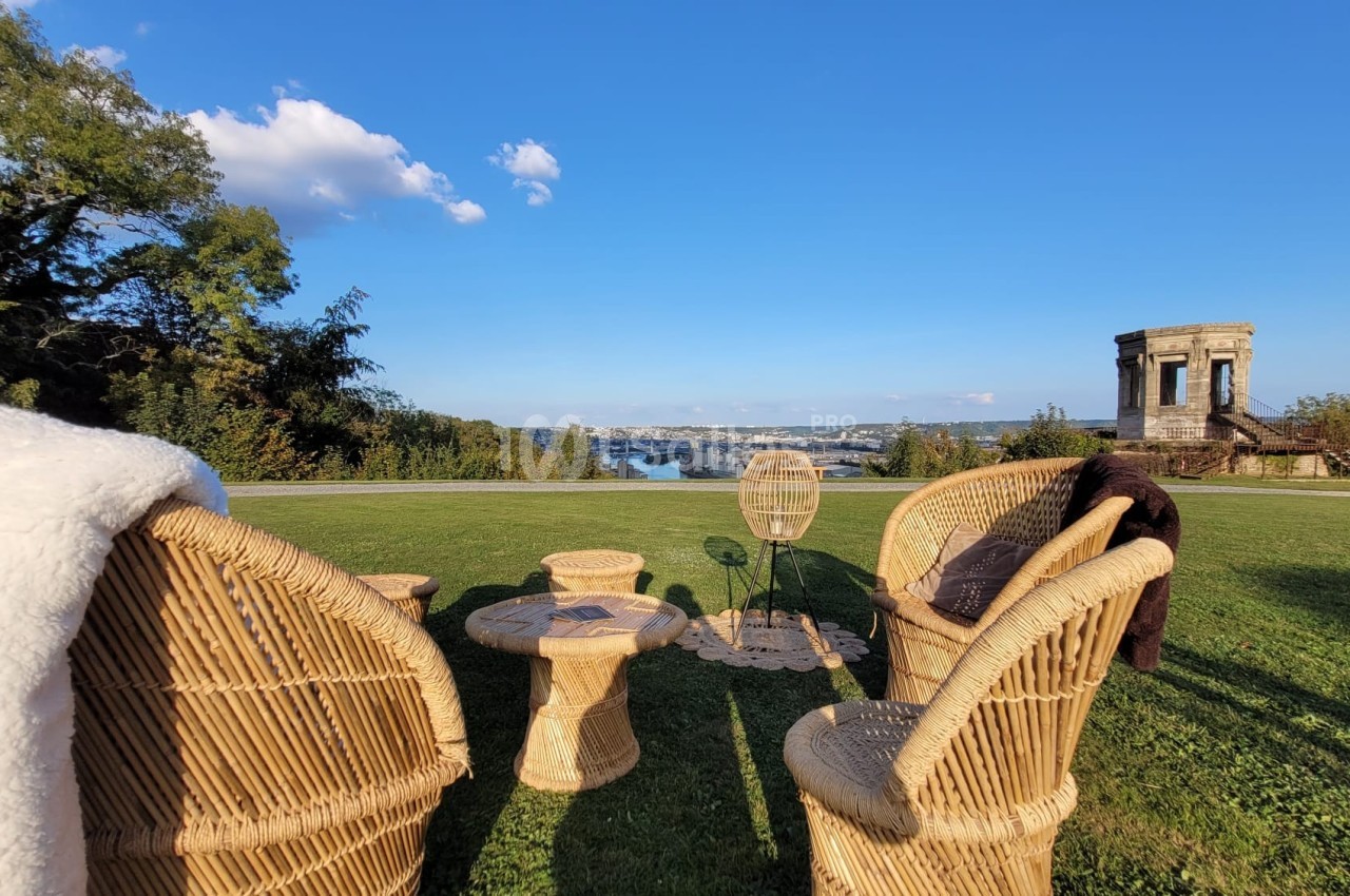 Chaises et table en rotin disposées sur une pelouse avec vue sur un fleuve et un bâtiment ancien à l'arrière-plan.