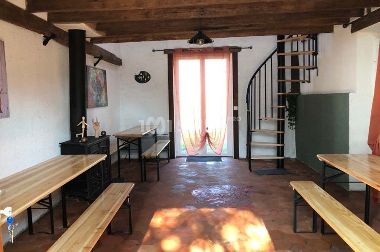 Salle lumineuse avec tables en bois, bancs, escalier en colimaçon, poutres apparentes et porte vitrée donnant sur l…