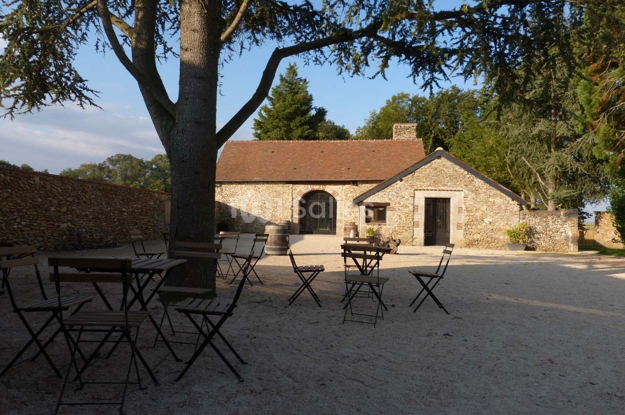 Cour extérieure avec des tables et chaises en métal, devant une maison en pierre entourée d'arbres et d'un mur.