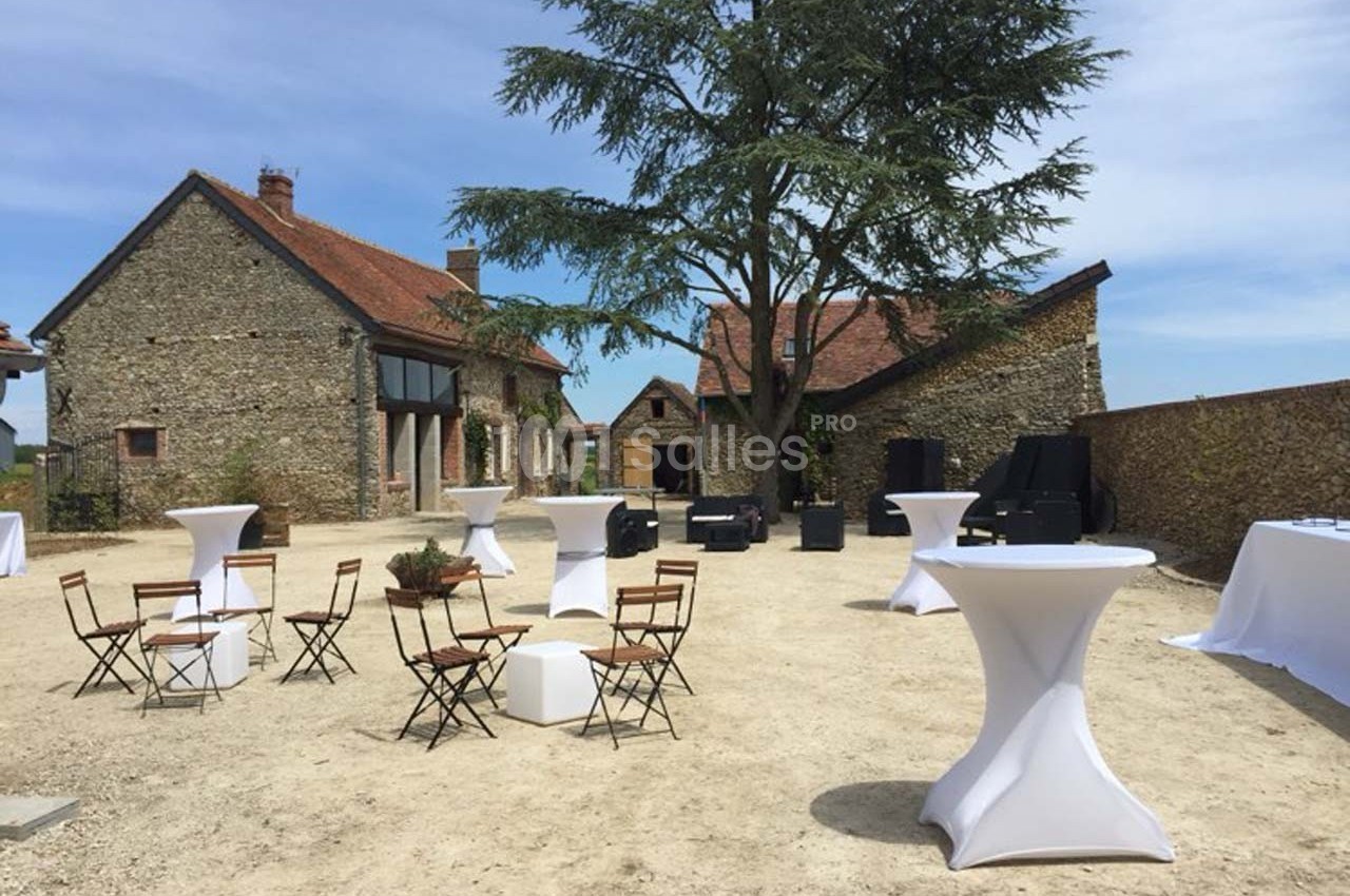 Cour extérieure avec tables hautes et chaises pliantes disposées devant des bâtiments en pierre sous un grand arbre.