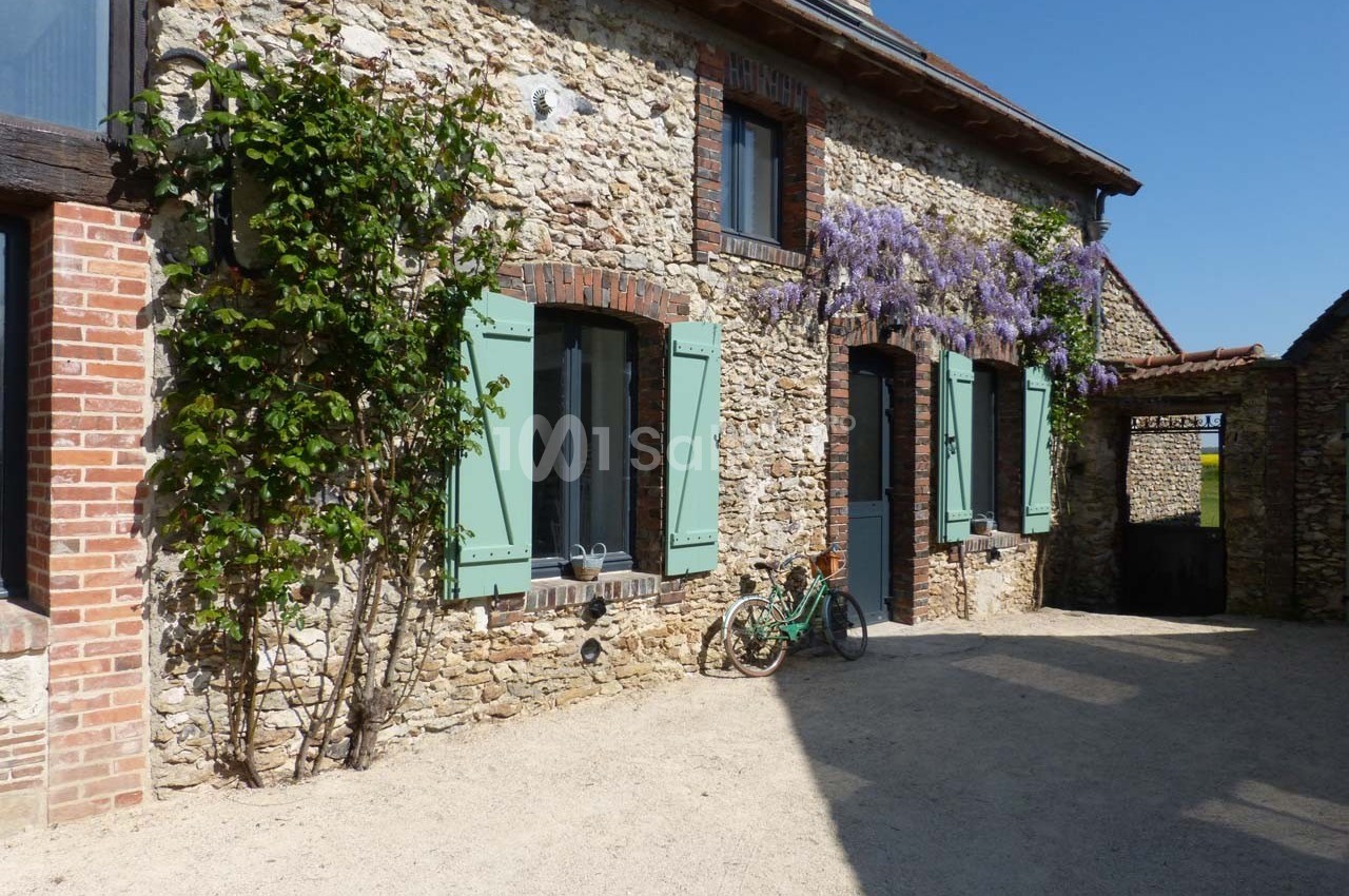 Façade en pierre d'une maison avec volets verts, glycine en fleurs et une bicyclette appuyée contre le mur.
