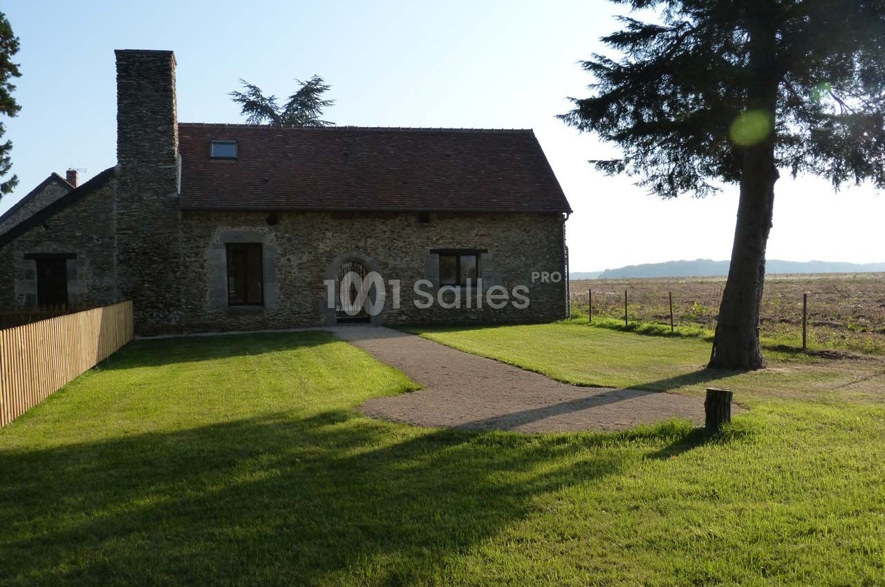 Maison en pierre avec toit en tuiles, entourée d'une pelouse et d'arbres, donnant sur un champ en arrière-plan.