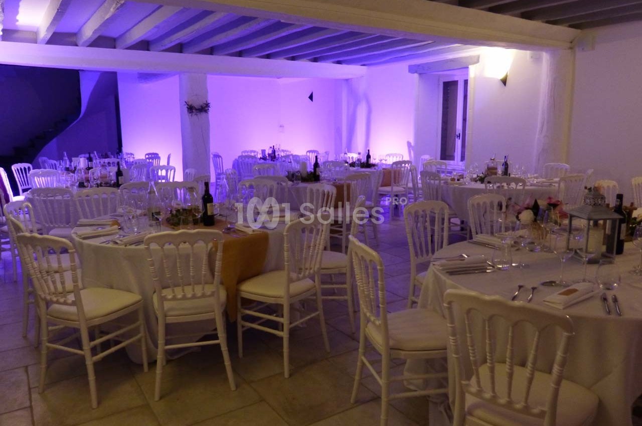 Salle de réception décorée avec des tables rondes dressées, chaises blanches et éclairage violet tamisé.