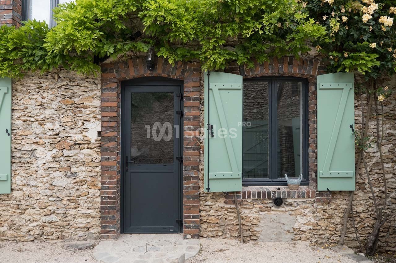 Façade en pierre avec porte noire, fenêtre à volets verts ouverts et végétation grimpante au-dessus.