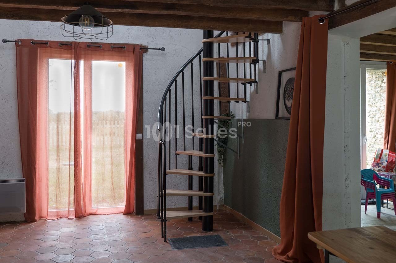 Escalier en colimaçon noir dans une pièce lumineuse avec sol en terre cuite, rideaux orange et poutres apparentes.