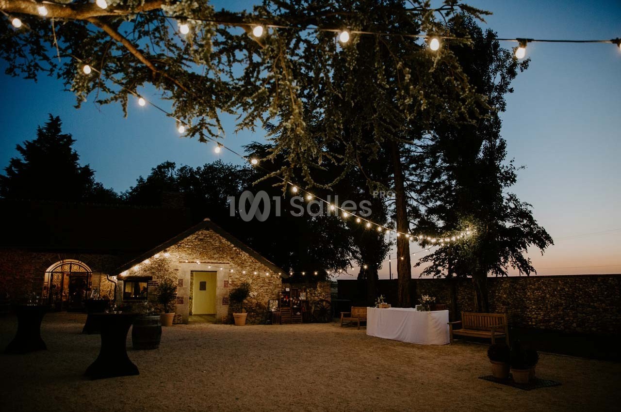 Cour extérieure éclairée par des guirlandes lumineuses au crépuscule, avec une maison en pierre et des tables décorées.