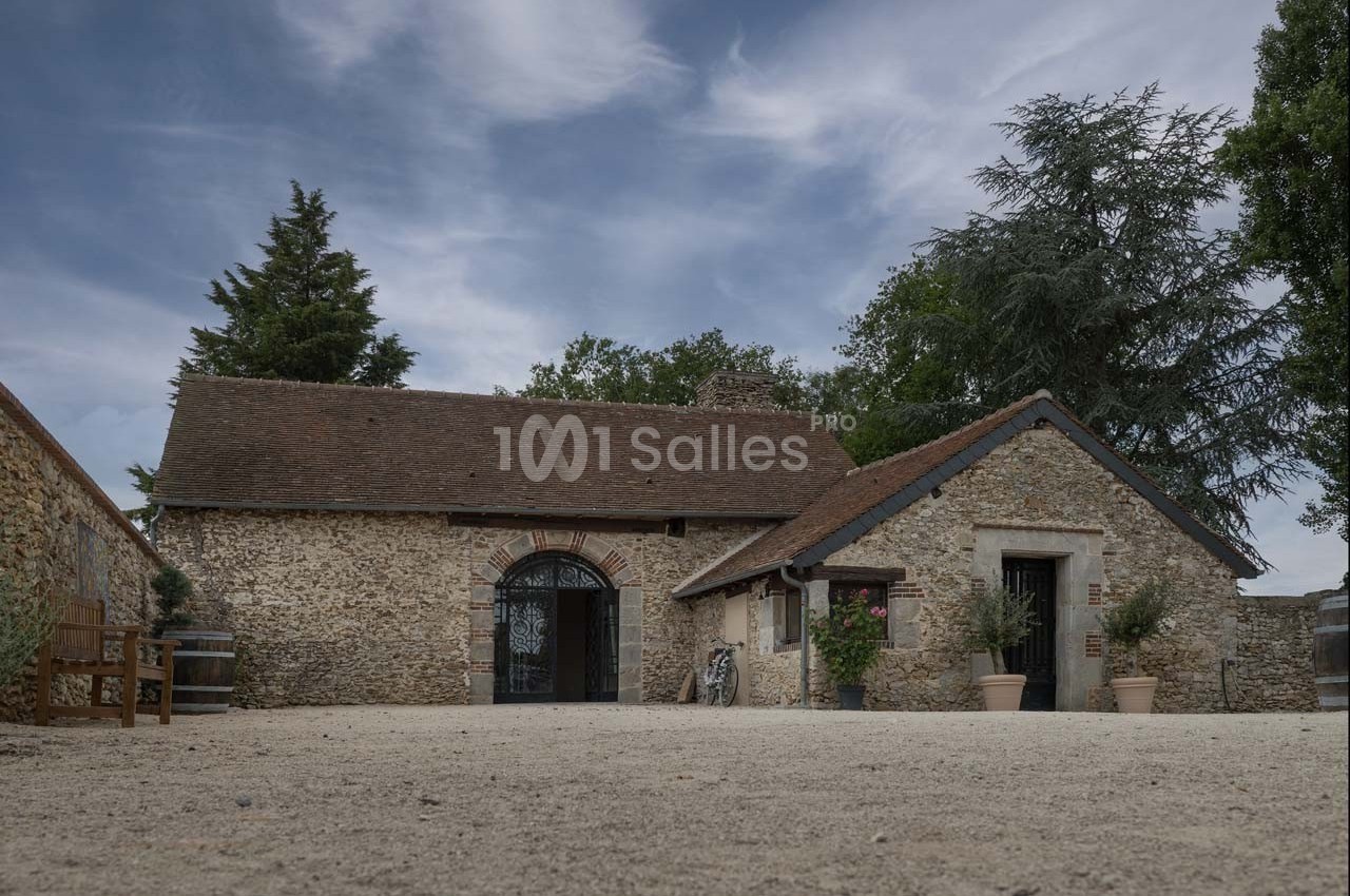 Bâtiment en pierre avec toit en tuiles, entouré d'arbres, sur une cour en gravier sous un ciel partiellement nuageux.