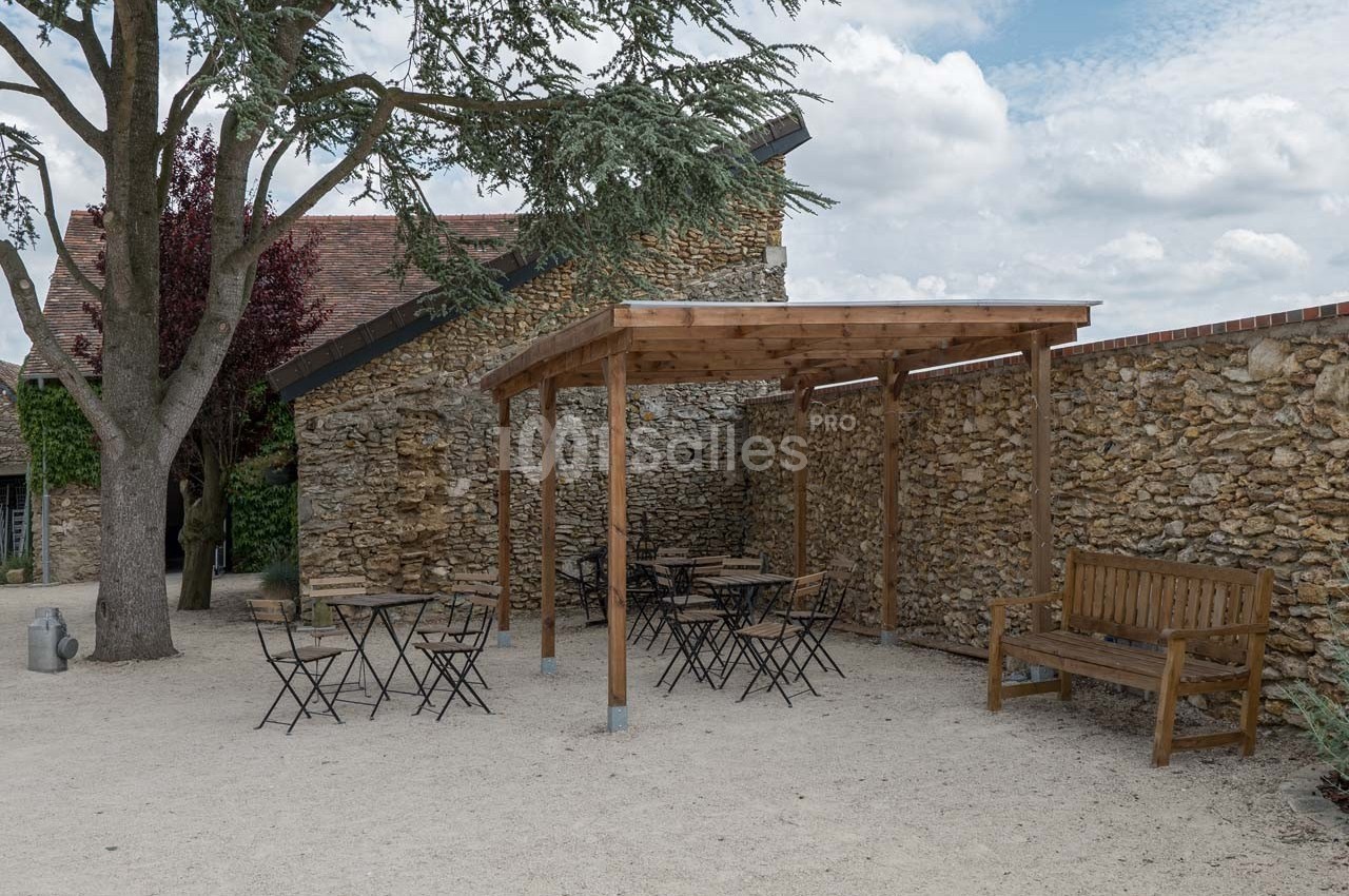 Terrasse extérieure avec pergola en bois, tables et chaises sur sol gravillonné, près d'un mur en pierre et d'un arbre.