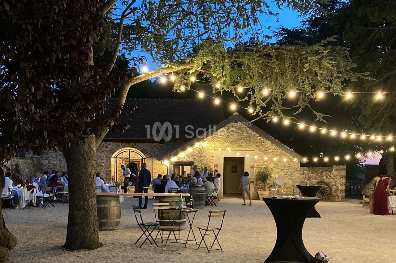 Cour extérieure éclairée par des guirlandes lumineuses, avec des tables et des invités rassemblés près d'un bâtiment en…