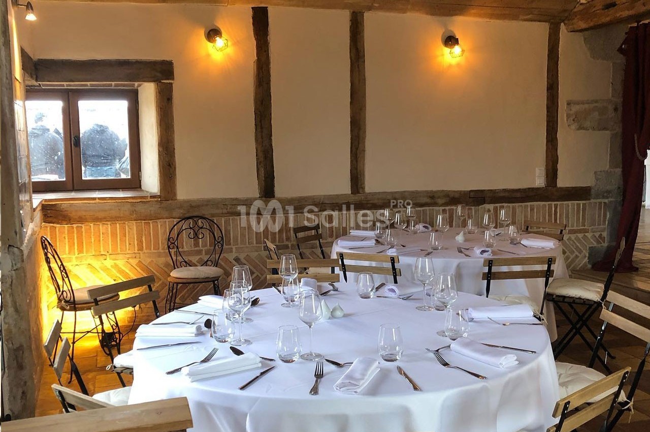 Salle rustique avec murs en bois et briques, tables rondes dressées avec nappes blanches et chaises en métal.