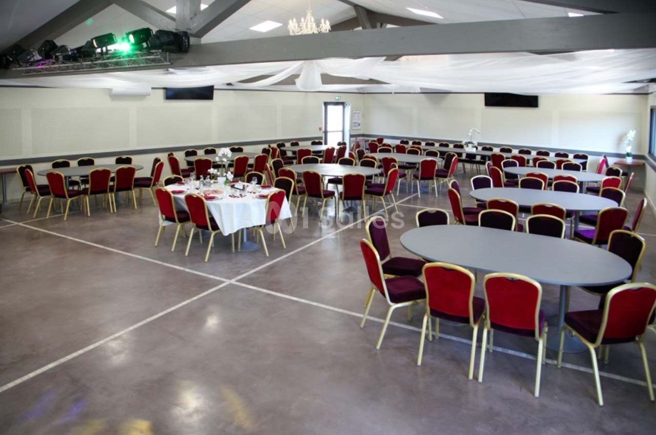 Salle de réception avec tables rondes et chaises rouges, décor sobre et plafond avec poutres apparentes.