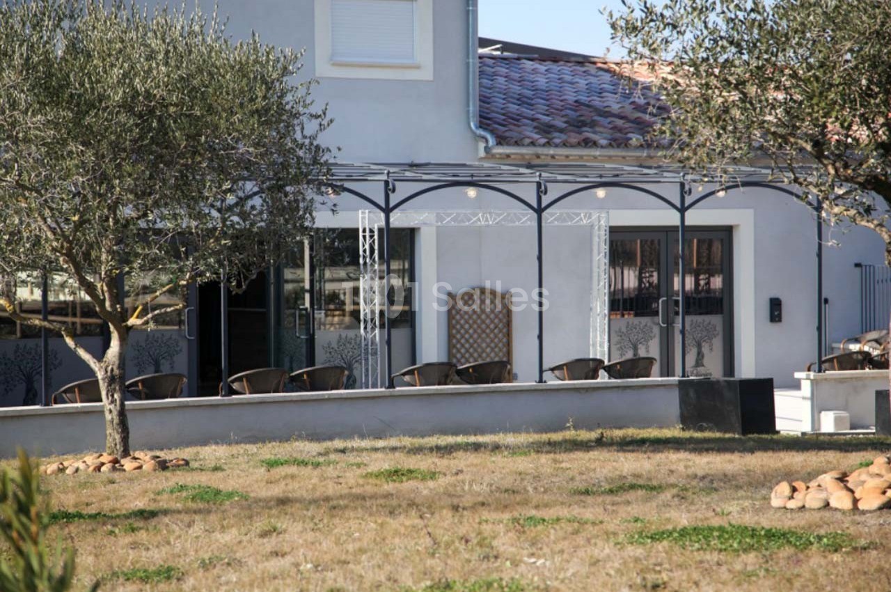 Façade d'une maison avec terrasse couverte, chaises en rotin, arbres et pelouse sèche au premier plan.