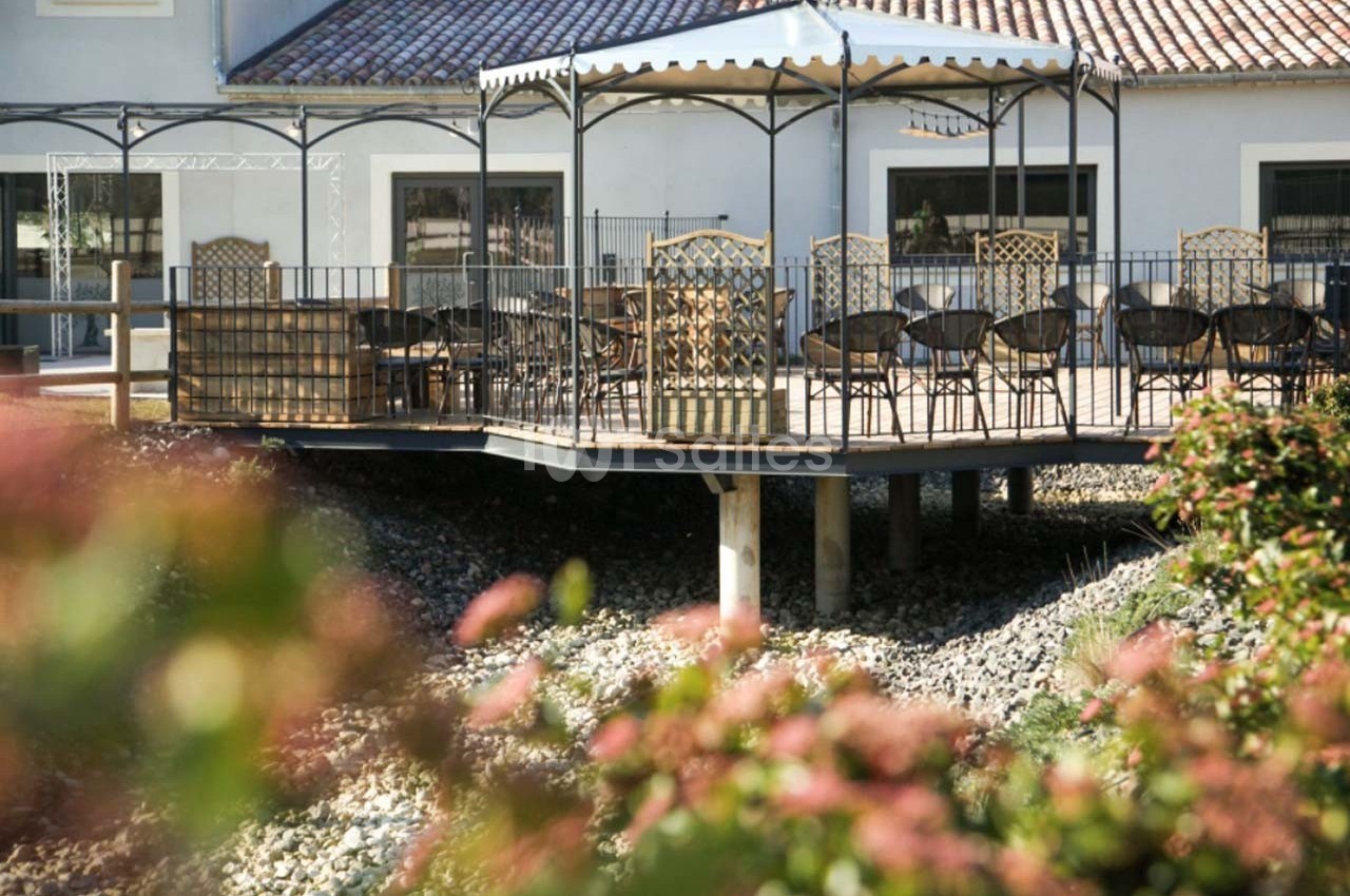 Terrasse surélevée avec pergola, équipée de tables et chaises, entourée de végétation et située devant un bâtiment clair.