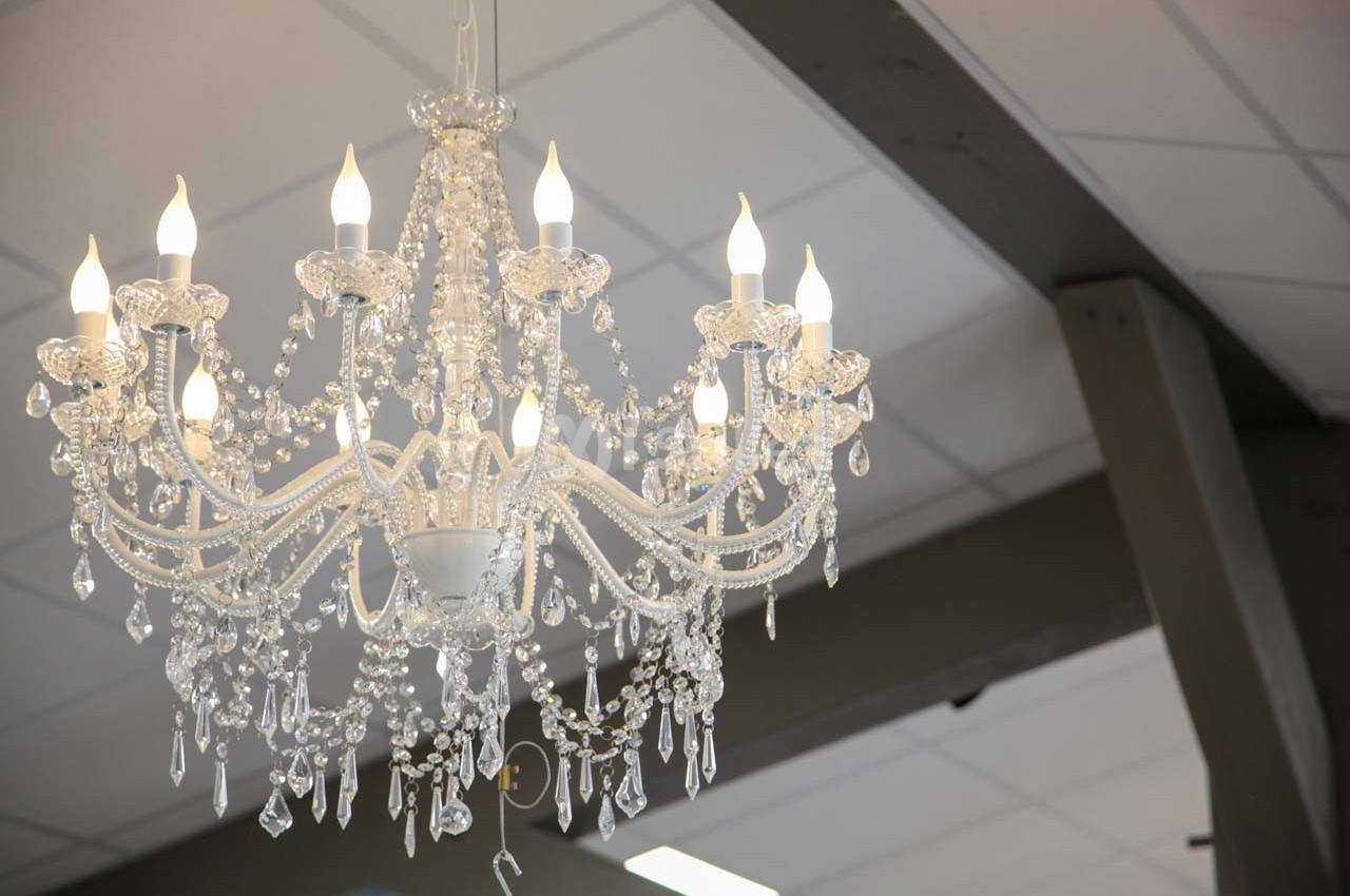 Lustre en cristal suspendu, allumé, avec des ampoules en forme de bougie, dans une pièce au plafond blanc et poutres grises.