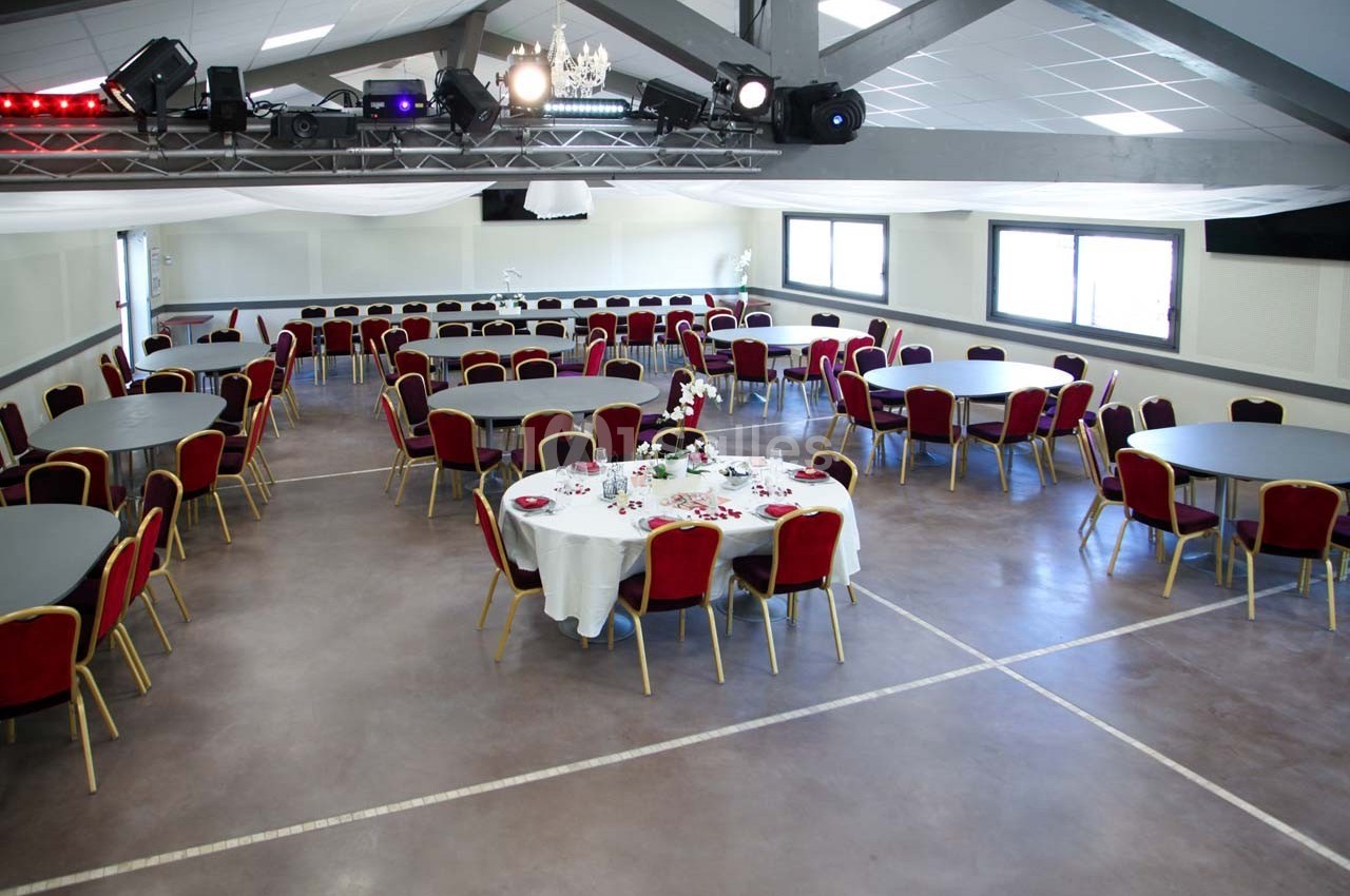 Salle de réception avec tables rondes dressées, chaises rouges et éclairage naturel provenant des fenêtres.
