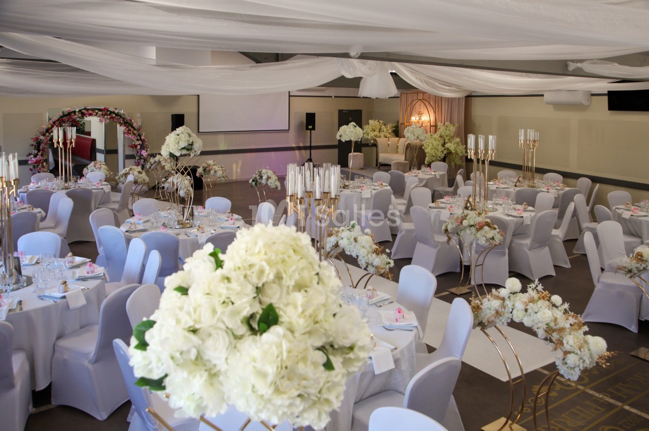 Salle de réception décorée avec des fleurs blanches, des arches florales et des tables dressées pour un événement.