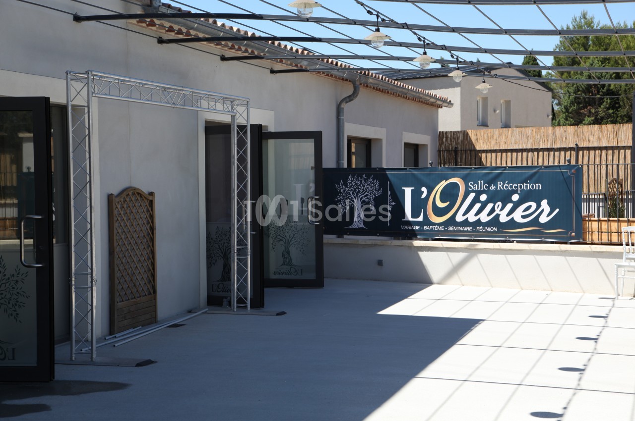 Entrée d'une salle de réception avec une bannière ’L'Olivier’ et des portes ouvertes donnant sur une terrasse.