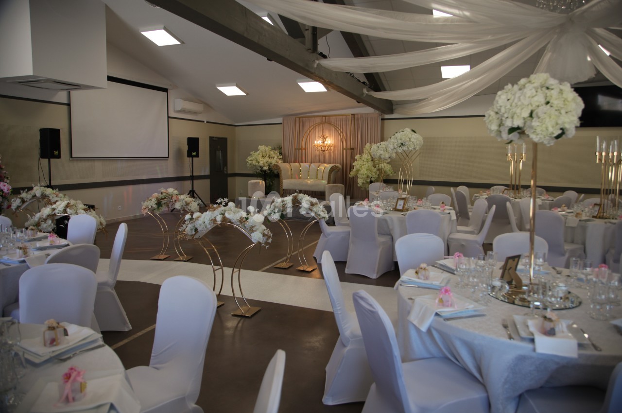 Salle de réception décorée avec des tables rondes, nappes blanches, compositions florales et un espace scénique éclairé.