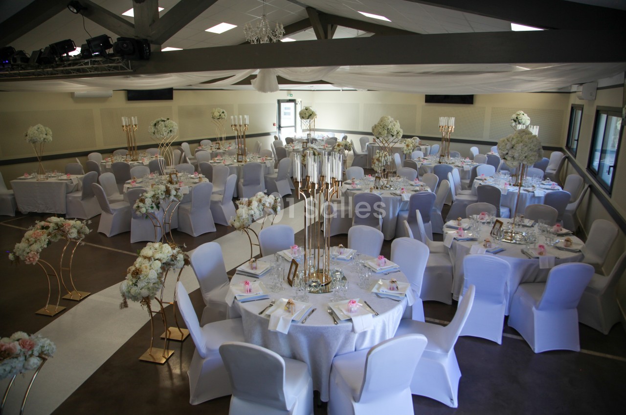 Salle de réception décorée pour un mariage avec des tables rondes, nappes blanches, chandeliers et arrangements floraux.