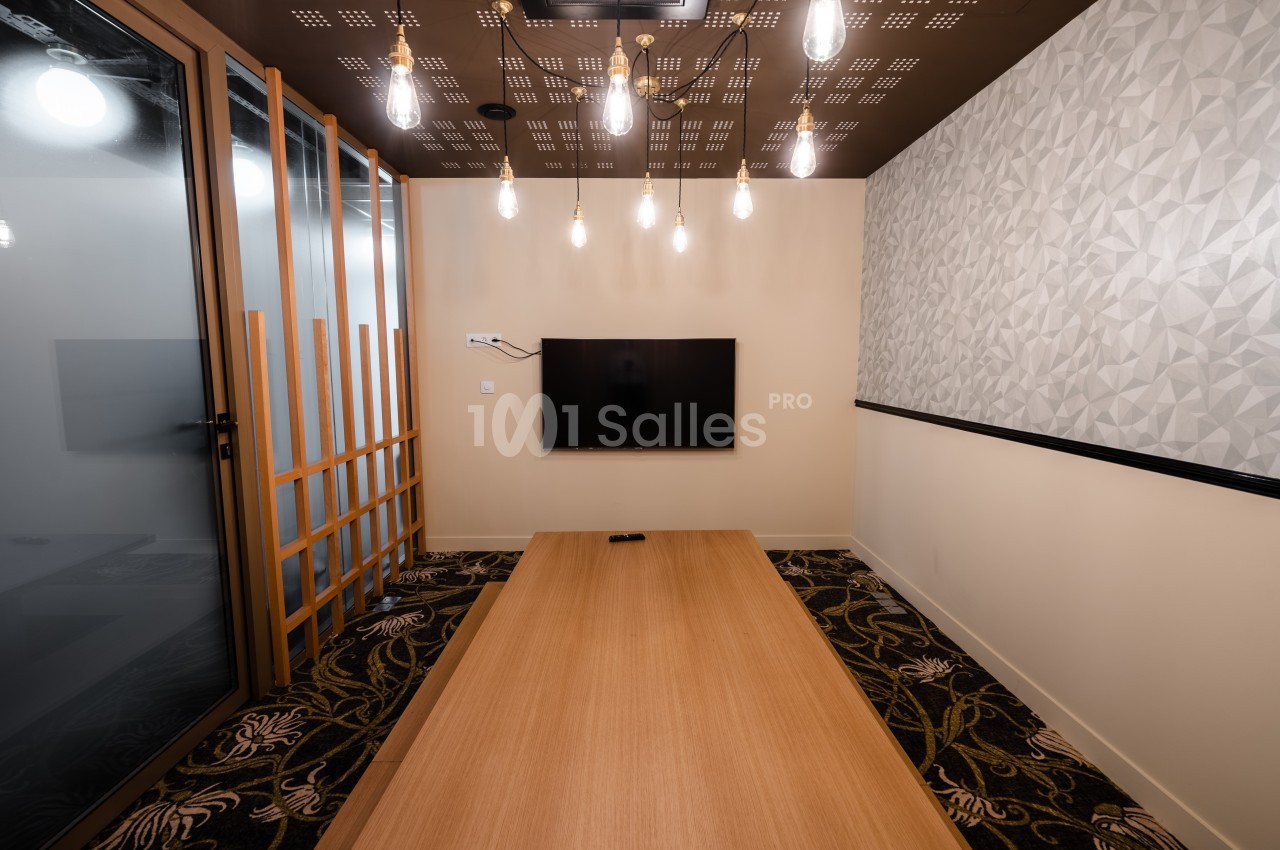 Salle de réunion moderne avec table en bois, écran mural, éclairage suspendu et cloison vitrée.